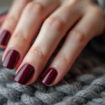 Mains féminines aux ongles courts avec gel bordeaux sur fond de laine grise en hiver, sans décollement ni casse